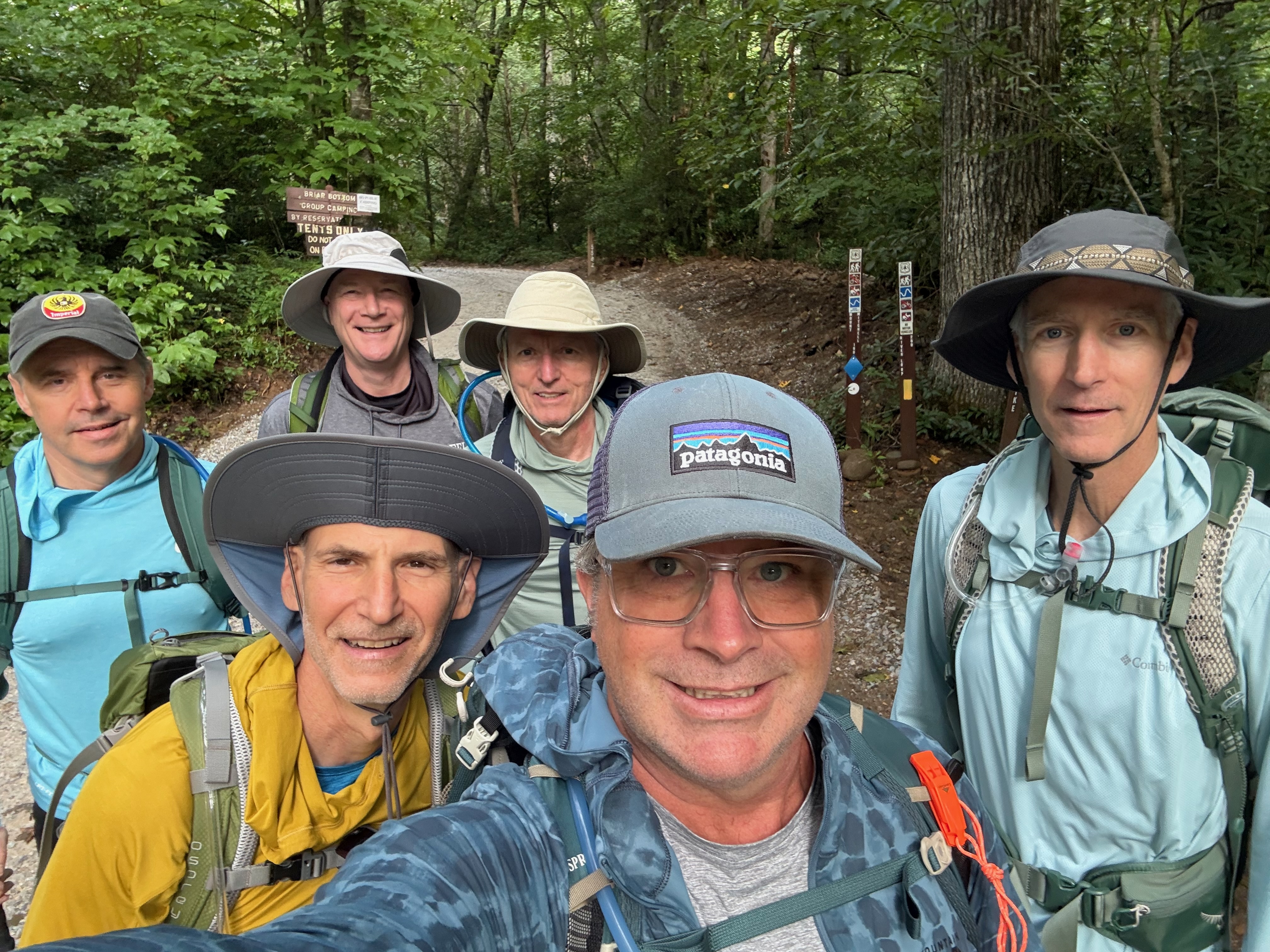 A group of six men smiling for a selfie while hiking in a wooded area, wearing outdoor gear and hats, with trail signs visible in the background.