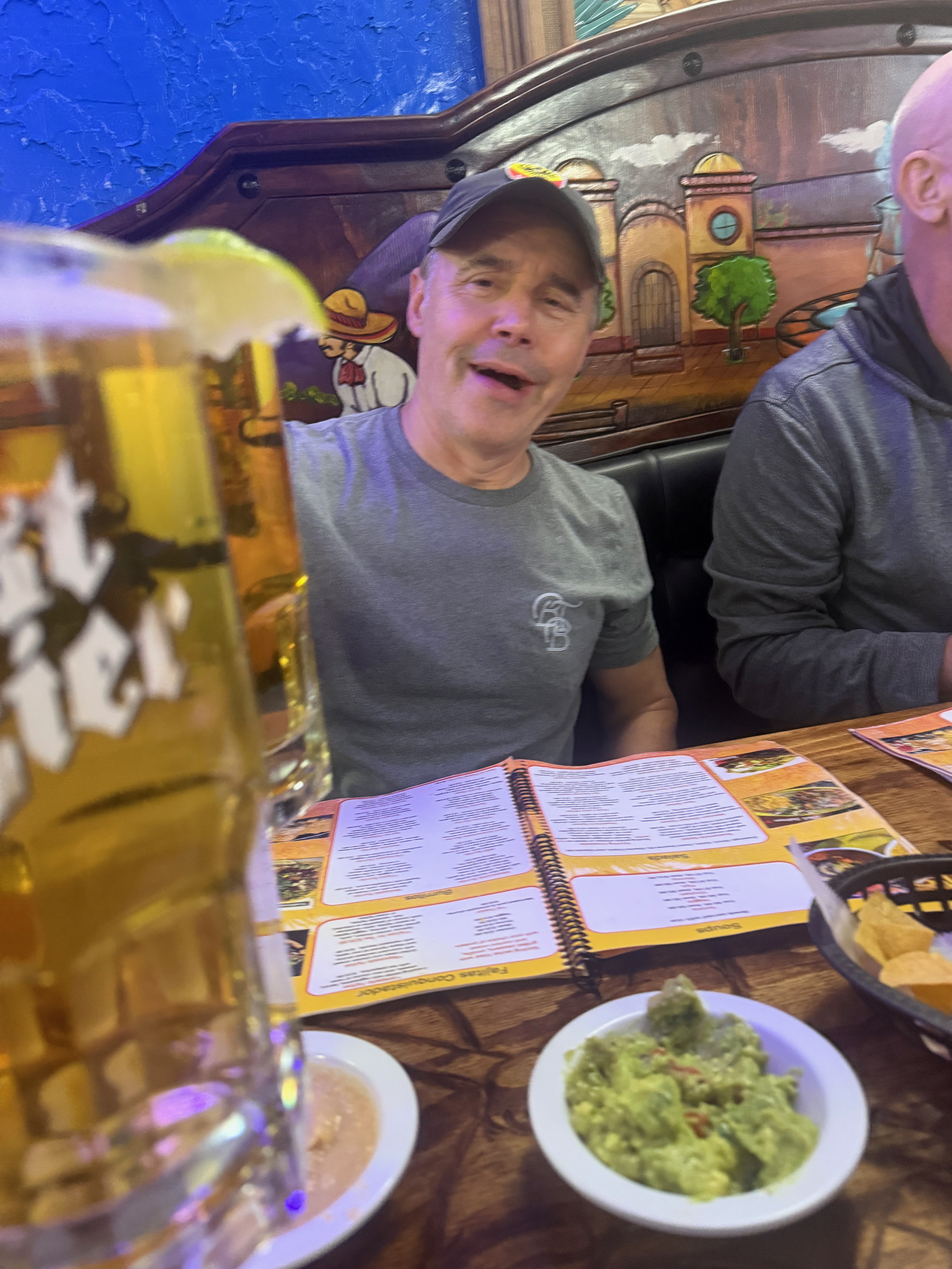 A man smiling at a table with a beverage and food items, including guacamole and a menu, in a restaurant setting.