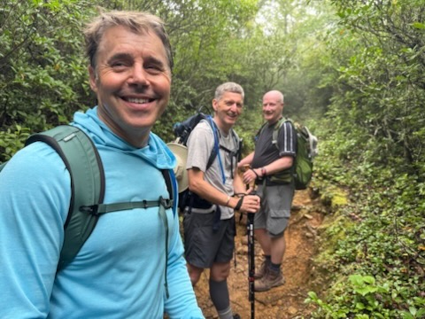 Three men smiling while hiking on a forest trail surrounded by lush greenery.