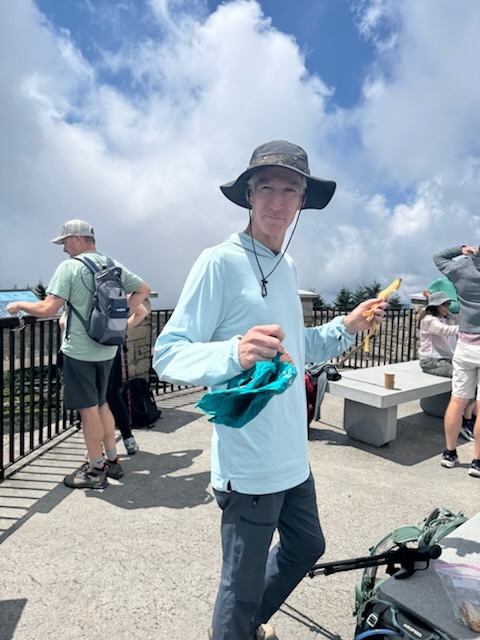 A man wearing a blue long-sleeve shirt and a wide-brimmed hat stands outdoors, holding a yellow object in one hand and a teal cloth in the other. He appears to be at a scenic lookout with other hikers in the background.