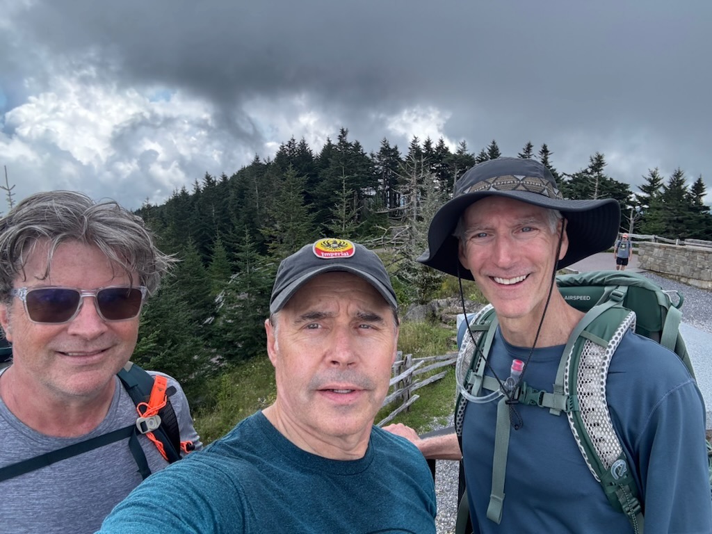 Three men posing for a selfie outdoors on a mountain trail surrounded by trees and cloudy skies.